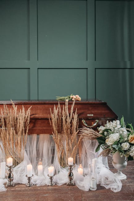 A floral arrangement featuring white and pale peach roses, white lilies, and greenery arranged in a white vase placed on a wooden surface. Surrounding the vase are tall, dried pampas grasses in shades