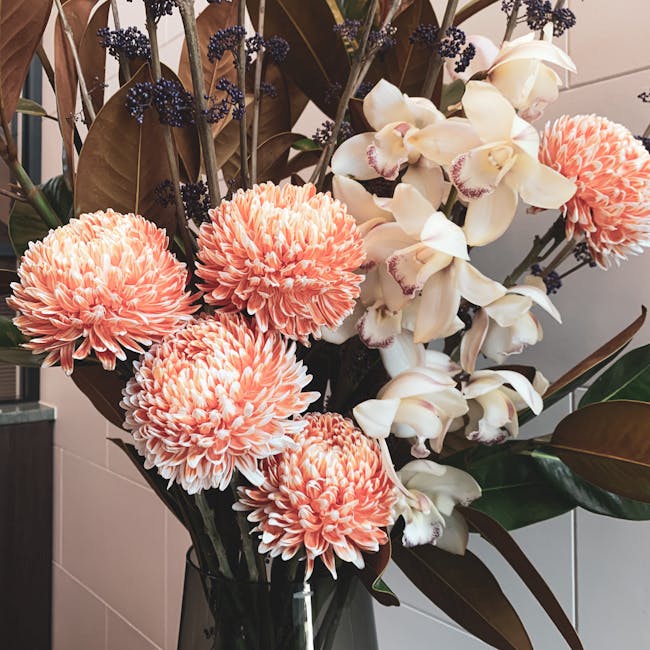 A tall, elegant floral arrangement in a black glass vase featuring large, round peach-colored chrysanthemums with densely petaled blooms, accented by creamy white orchids with pink-rimmed petals, and 