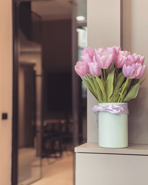 A pastel green cylindrical vase with a lavender ribbon tied around its neck contains a bouquet of fresh pink tulips with vibrant green stems and leaves. The bouquet is arranged loosely with the tulips
