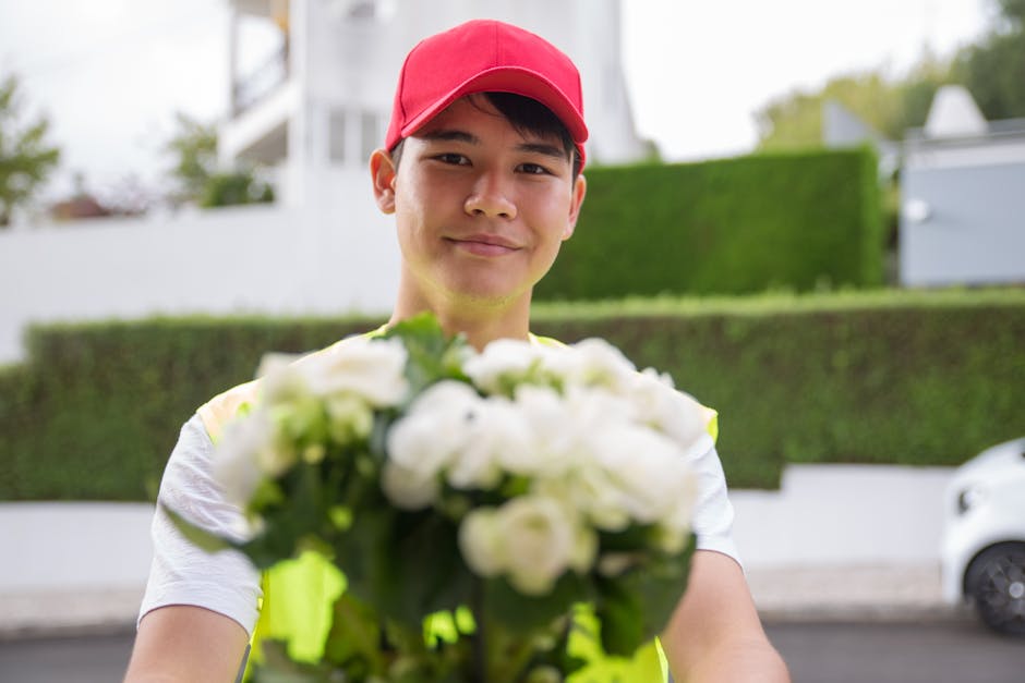 A delivery person wearing a red cap and a high-visibility vest is smiling slightly while holding a bouquet of fresh white flowers, which include roses and possibly lisianthus, wrapped in green foliage