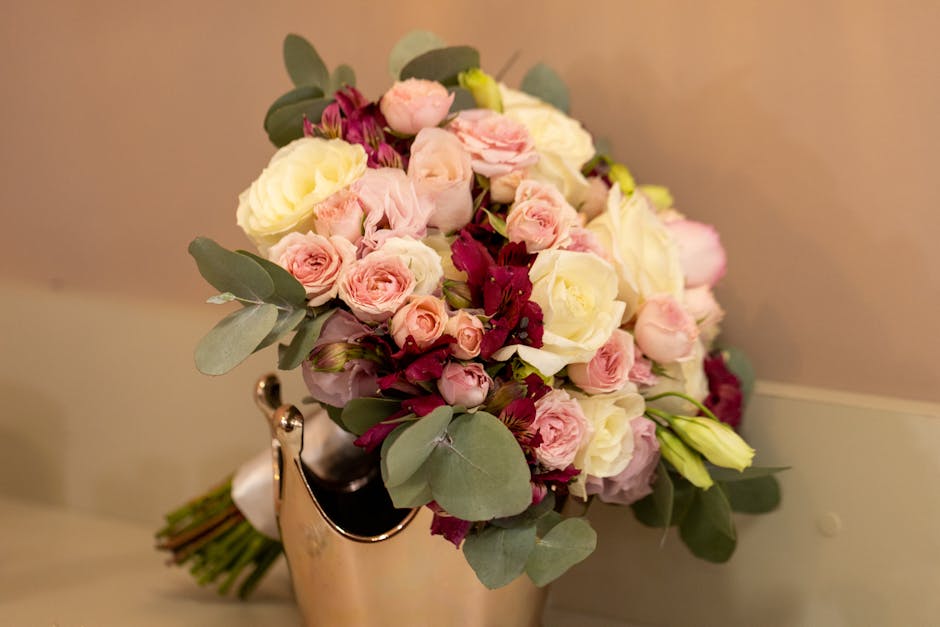A floral bouquet arrangement featuring pale pink and cream roses, complemented by deep burgundy flowers and soft green eucalyptus leaves. The bouquet is hand-tied with visible stems wrapped in white r