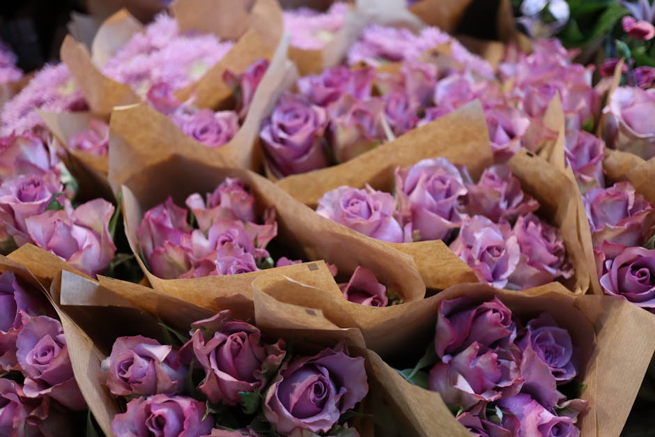 Close-up of multiple small bouquets of fresh lavender-colored roses wrapped in light brown kraft paper, arranged closely together on a surface. The roses have delicate, slightly ruffled petals with a 