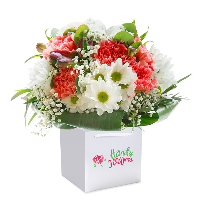 An exquisitely crafted bouquet from Grove Park is displayed upright in a classic white square gift bag, poised against a spotless white backdrop that lets every hue and texture shine. The arrangement brims with soft coral and peach carnations, each bloom laden with elegantly ruffled petals that lend a tactile richness and visual depth. Among them nestle pure white daisies with vivid yellow-green centers, pouring cheerful brightness into the composition and channeling the airy parkscapes of Grove Park and Chiswick. Dainty sprays of baby's breath spill lightly through the display, their presence adding a gentle, almost weightless counterpoint. Tucked on one side, closed purple-tipped buds introduce subtle intrigue and promise, hinting at the unfolding of future celebrations. Deep, luxurious green foliage forms a natural, glossy frame at the bouquet's base, much like the lush woods that line the riverside paths near Strand-on-the-Green. Every bloom is expertly placed for harmonious fullness, while the white ribbon handles of the gift bag suggest care and readiness for delivery across Grove Park or for hand-off along Staveley Road. Rendered in clear, even light, the arrangement presents a sense of abundance and quiet charm-an impeccable choice for birthdays, milestones, or acts of affection. Local floral skill and Grove Park's signature warmth are woven through each petal and leaf.