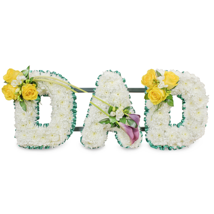 A thoughtfully composed sympathy tribute that spells out DAD in large, freestanding block letters, each letter entirely covered with a thick layer of pristine white chrysanthemums so the surface appears soft and uniform like a white blanket of blooms. The outer and inner rims of every letter are neatly edged with a ruffled, dark green satin ribbon, giving a crisp outline that sets the white blooms apart from the pale background and echoes the trimmed foliage often used in formal memorial work. Visible behind the letters are slim, dark green metal connecting bars that run horizontally, a reminder of the hidden support structure that keeps the letters aligned and dignified for display on an easel or memorial stand. Floral accents are placed with care: the left D hosts a compact cluster on its upper left curve made up of three to four bright yellow roses, small white freesia-like fillers, and fresh green leaves, while two long slender green stems arch diagonally toward the central A to create a sense of movement. The A itself is decorated on its lower left diagonal with two deep purple-to-burgundy calla lilies; their smooth, sculptural forms provide a quiet, contemplative centre, surrounded by tiny white accents. The right D mirrors the left with the same yellow rose cluster on its upper right curve, completing the visual symmetry. Photographed in soft, neutral light against a plain white ground, this arrangement conveys calm and respect - a classic choice for funerals or wakes in Grove Park and surrounding neighbourhoods, easily delivered to local churches and chapels with a restrained, comforting scent of fresh roses and green leaves.