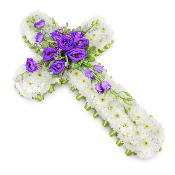 A detailed funeral tribute in the form of a Christian cross is shown resting on a bright white background, the clarity of the setting drawing full attention to the workmanship of the flowers. The cross itself is completely covered with small white chrysanthemums, so tightly arranged that their petals form a continuous, cushion-like surface. The blooms are fresh and luminous, with light green centres that punctuate the white and give the design a subtle, natural rhythm. Around the edges, a crisp line of lime-green ribbon wraps the form, neatly finishing the sides and defining the silhouette with a gentle flash of colour that remains soft and respectful. At the central junction where the arms of the cross meet, a lush gathering of deep purple lisianthus forms a striking focal point. The lisianthus blossoms are plush and ruffled, their petals curling around each other in layers that evoke both luxury and serenity. Interspersed are small, unopened buds, slender leaves, and fine filler flowers, which lend height, depth, and a touch of airy movement. From this central spray, a small cascade of lisianthus blooms and buds extends diagonally down one side of the cross, breaking the strict symmetry just enough to create an elegant, flowing line. The photograph is taken from a slightly overhead angle, bathed in soft, even light that reveals every nuance of texture, while a delicate shadow beneath the cross gives it presence, as though laid gently on a table in a Grove Park church, local funeral parlour, or family home near the station. The combination of pure white and earnest purple creates a tribute suited to memorial services, church funerals, and graveside ceremonies in Grove Park, conveying heartfelt condolence, faith, and lasting remembrance.